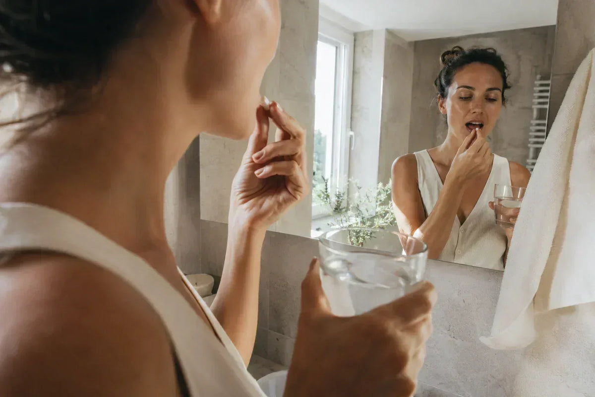 A woman is standing in front of the mirror in the bathroom, putting zinc in her mouth and holding a glass of water